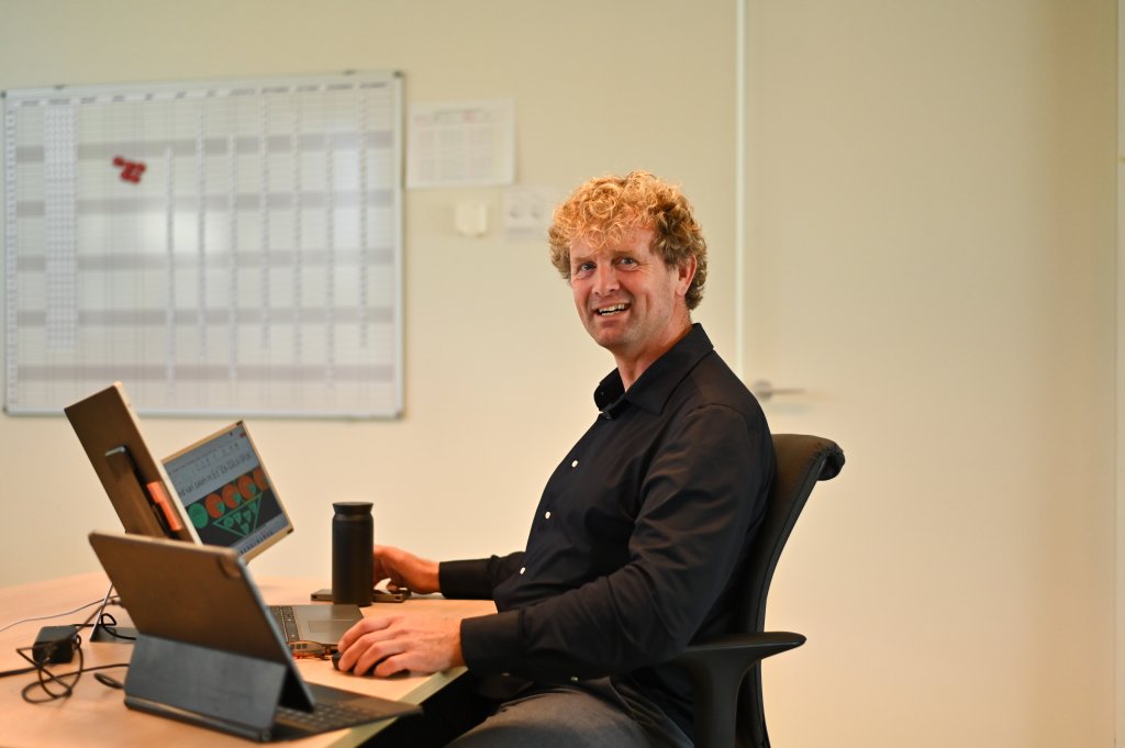 Edzart zit aan tafel achter zijn laptop op de werkvloer en kijkt opzij in de camera. Achter hem hangt een whiteboard met de planning.